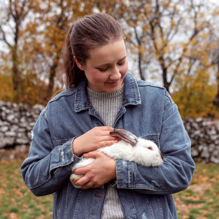 Femme souriante tient un lapin blanc dans ses bras, entourée d'un paysage automnal.